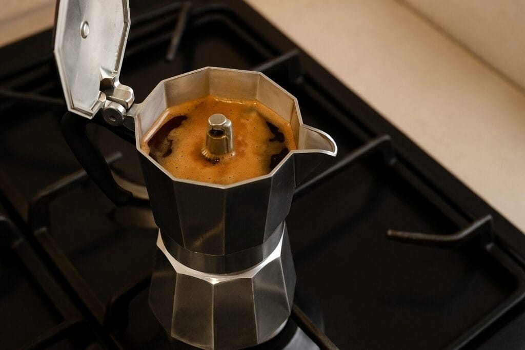 A stainless steel Moka pot brewing coffee on a gas stove, with fresh espresso bubbling in the upper chamber